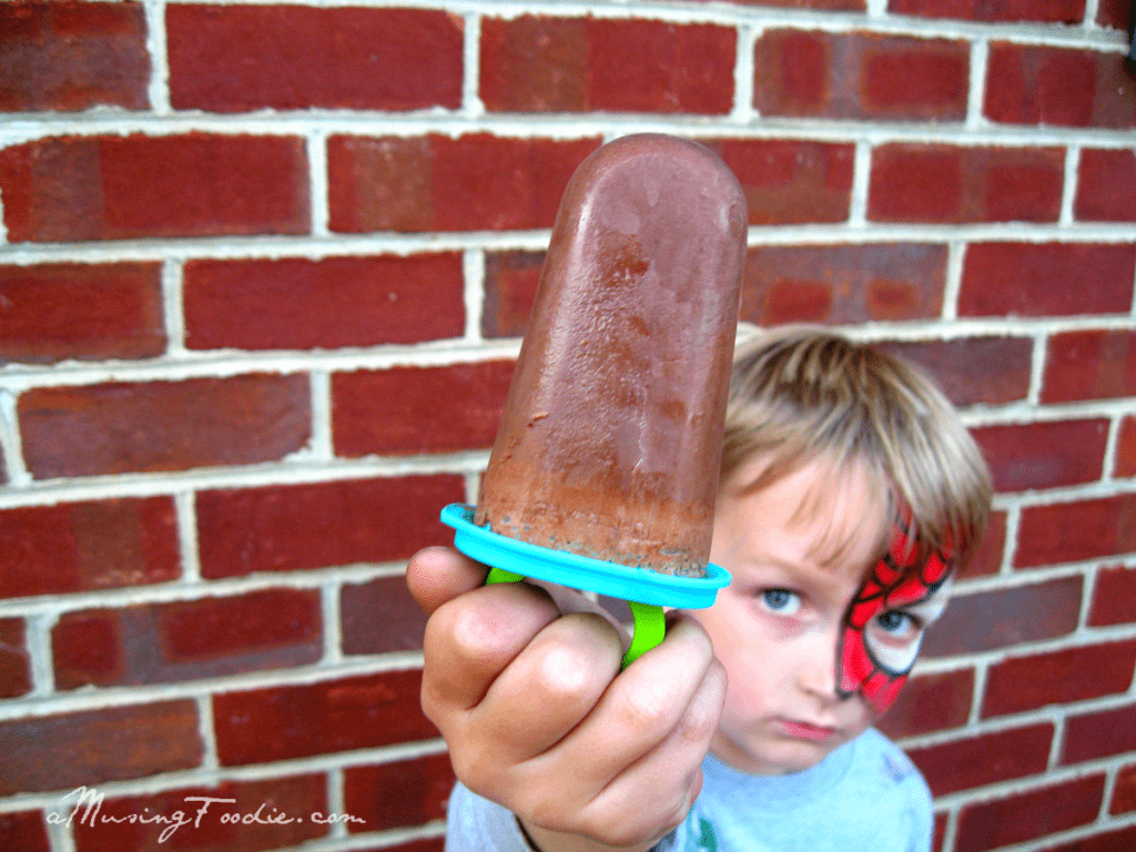 Boy in Spider-Man face paint enjoys a chocolate popsicle against a rustic brick wall.